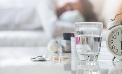 Closeup glass of drink water and pills on white table with  blurred background of man sleeping on sofa, medicine and health care concept, copy space.