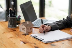 Cropped image of woman hand holding pen writing on blank paper with house model and keys on wooden table, real estate business concept.