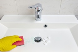 A hand in yellow protective gloves cleans the sink in the bathroom with a sponge and a tablet bleach cleaner for sanitary equipment in the bathroom. Cleaning and cleaning the washbasin