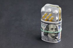 Pills and tablets in blister packs are wrapped with dollar bills fastened with a rubber band on a black concrete background. Rising prices for medicines