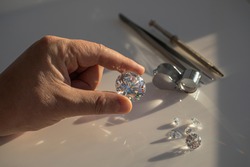 Close-up of the hand of jeweller gemologist with large size round cut diamond on desktop with magnifier and tweezers. Buyer diamond expert checking color of diamond in natural light.
