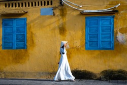 Vietnamese woman wearing a white ao dai or traditional tunic and pant suit and a non la or conical straw hat while walking in front of a gold stucco building with brilliant turquoise shuttered windows
