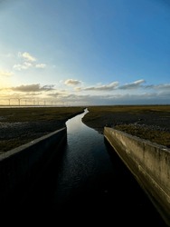 A serene water channel meanders through a marsh under a dusky sky, with wind turbines on the horizon reflecting the sustainable synergy between land and technology.