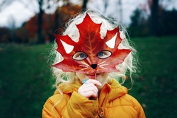 Portrait of cute child kid with blue eyes holding autumn fall red maple leaf painted mask in front face. Girl playing pretending to be fox cat. Fairy dreamy authentic childhood. Halloween holiday.