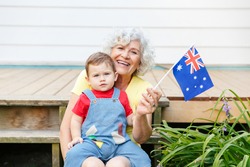 Happy old woman grandmother with grandson baby holding Australian flag. Smiling old lady with kid child on home backyard waving Australia flag. Family citizens celebrating Australia Day.