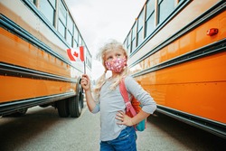 Caucasian girl student with face mask holding Canadian flag. Student kid near yellow school bus in Canada. Education and back to school in September. New normal during coronavirus. 