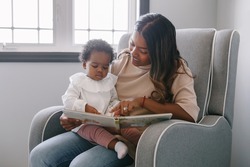Mixed race Indian mom reading book with African black baby girl toddler at home. Early age children education development. Family authentic candid lifestyle. 