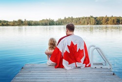 Father and daughter wrapped in large Canadian flag sitting on wooden pier by lake. Canada Day celebration outdoor. Dad and child sitting together on 1 of July celebrating national Canada Day.
