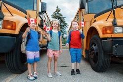 Proud happy children students boys and girl holding waving Canadian flags. Education and back to school in September. Group of friends kids near yellow school bus outdoors on school yard. 