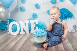 Portrait of cute adorable blond Caucasian baby boy with blue eyes in jeans overall celebrating his first birthday with gourmet cake letters one and balloons cake smash in studio
