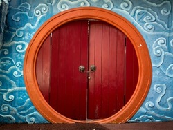 Round shaped door with orange frame and red wooden planks, surrounded by blue cloud sculpture. Traditional Chinese lion head door knocker.