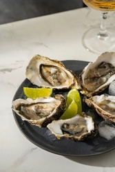 Fresh oysters with lime, onion on a grey round plate. Oyster season. Macro seafood dish. Oysters on the half shell with glass of sparkling wine on white table, beautiful light. Healthy food concept.