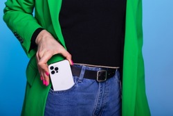 Young woman takes out or puts in a pocket mobile phone. Woman wears green blazer and blue jeans. Smartphone with three cameras in front pocket on blue background. Mockup for print or design template