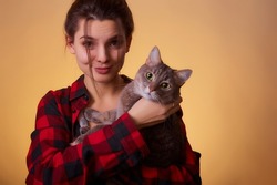 Closeup portrait of woman in checkered shirt posing on yellow background, with cat in hands. Photoshoot with a pet. Lady holds a cat near her face and funny laughing