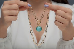 Gold necklace with blue stone on hand of lady with blue nail polish in white clothes.