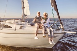 Happy senior couple sitting on the side of sail boat or yacht deck floating in sea. Man and woman drinking wine or champagne and laughing
