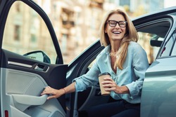 Happy attractive woman or business lady wearing eyeglasses holding cup of coffee and getting out of her modern car
