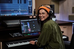 Portrait of beautiful young brunette, female artist smiling at camera while playing keyboard synthesizer, sitting in recording studio