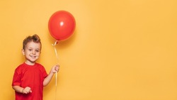 A happy Caucasian boy isolated on a bright yellow background holds a red balloon in his hands. A place for your text or advertisement.