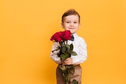 Boy in a white shirt, pants and a bow tie on a yellow background holds in his hands bouquet a red roses. A gift for my mother, grandmother.