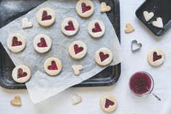 Flat lay image of heart shaped Linzer jam and jelly filled cookies. Baking treats concept including jam, cookie cutters, icing sugar and baking sheets in top down view. Romance and love food concept.