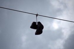 Silhouette of a tattered pair of high top athletic shoes that have been tossed over and are hanging from a utility wire against a darkening stormy sky.