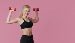 Fitness girl with perfect slim and fit body training muscles with dumbbells on pink background. Sporty woman in sportswear