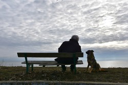 Lonely old man with dog. Senior man sitting on an old wooden bench and watching dark winter see, his dog quietly standing by. Nostalgic mood. 
