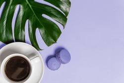 White cup of black coffee, two french macarons and green monstera leaf on purple background toned in trendy Very Peri color of the year 2022 from above. Top view, flat lay with copy space
