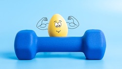 Funny Easter egg with cute face shows his muscles behind dumbbell on blue background. Strong athlete yellow egg character in theme of sports, training and healthy lifestyle. Protein and nutrition