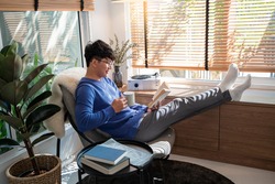 joyful young Asian man in casual clothes and eyeglasses reading book while relaxing at living room of his home