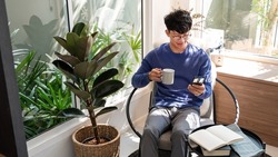 Handsome Asian teenager man wearing casual clothes in glasses using smart phone browsing Internet on mobile phone device at the living room 