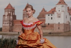 A young woman in an old orange dress walks on the shore of the lake near the castle
