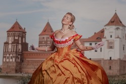 A young woman in an old orange dress walks on the shore of the lake near the castle