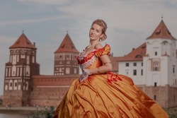 A young woman in an old fashion orange dress walks on the shore of the lake near the castle