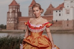 A young woman in an old fashion orange dress walks on the shore of the lake near the castle