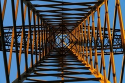 Steel structure of a high voltage pylon with wires, insulators seen from underneath in frog perspective with vanishing point and blue sky. Contrasting orange structure illuminated by sunset light