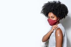 black teen girl shows the vaccine mark on her arm, wearing protective mask against covid-19, with a smile on her face, isolated on gray background