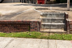 Sloping concrete parking lot ending at the top of a low brick retaining wall and cement block stairs to a sidewalk and grass, horizontal aspect