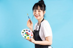 Beautiful Asian female painter holding quill pen and color palette on blue background
