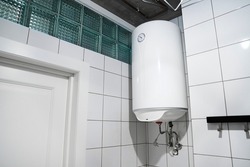 Household budget water heater hanging on the wall in boiler room. Modern gas tanked boiler in bathroom. Common electric storage tank water heater. Energy-efficient home heating system on white tiles