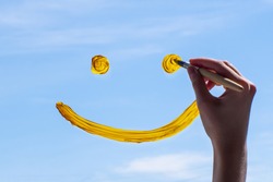 Close-up female hand draws a yellow smile on glass against a blue sky. The concept of joy for a new day, good luck