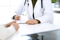Woman doctor and patient sitting and talking at medical examination at hospital office, close-up. Therapist filling up medication history records. Medicine and healthcare concept