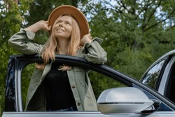 Blonde woman in hat staying next to car door. Young tourist explore local travel making candid real moments. True emotions expressions of getting away and refresh relax