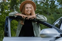 Blonde woman in hat staying next to car door. Young tourist explore local travel making candid real moments. True emotions expressions of getting away and refresh relax