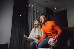 young cheerful family watching match on television and sit on comfortable brown sofa. Woman threw popcorn up in room. Screaming with joy in gray tone room. Wear colorful sweeter, blue jeans. 