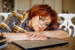 Tired mature lady in glasses lying down on laptop at desk in home room. Advanced pensioner practicing computer skills and learning modern technology. Online communication and distant future concept