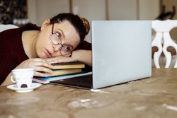 Tired young woman in glasses lying down on books and looking at laptop screen at desk in home room. Tiredness from monotonous study and computer overwork. Lack of energy motivation concept