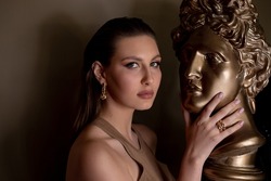 Portrait of young gorgeous woman with long dark hair, bright golden make-up, pink lips touching head face of bronze bust David by Michelangelo on grey background. Studio shot. Beauty, cosmetics, art