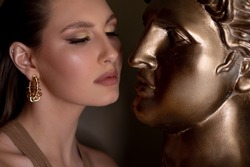 Portrait of young perfect woman with bright shining make-up, pink lips, wearing round golden earrings, looking at lips of bronze bust David by Michelangelo on grey background. Beauty, cosmetics, art